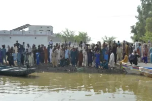 A large group of people standing beside floodwater in a village.