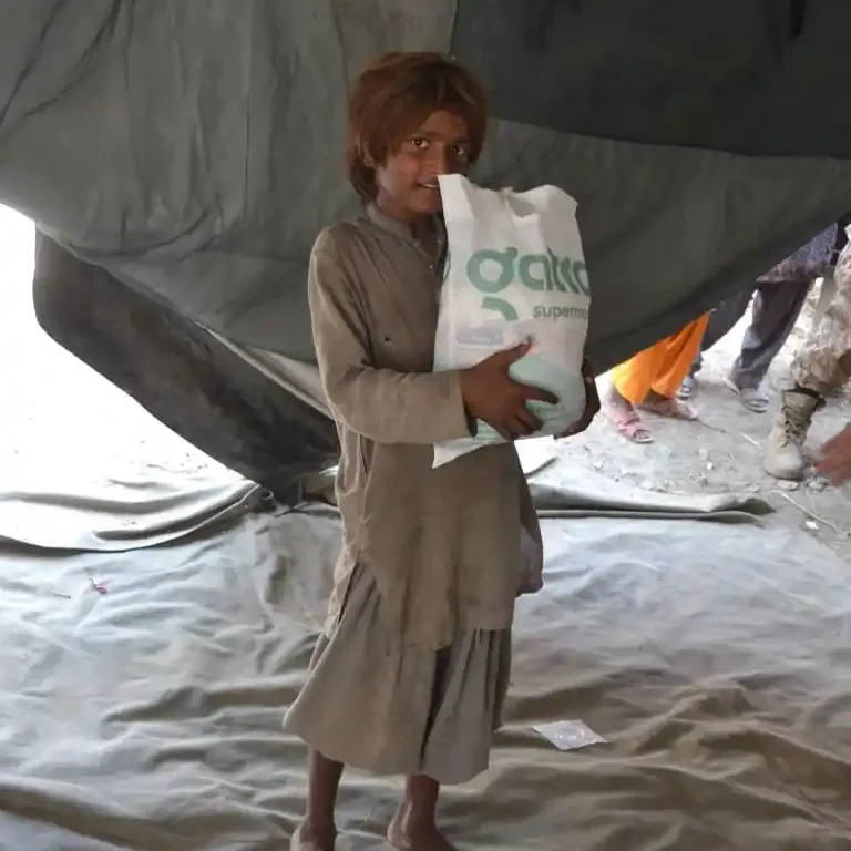 A resilient child impacted by floods holds a relief bag in a temporary Pakistani shelter.